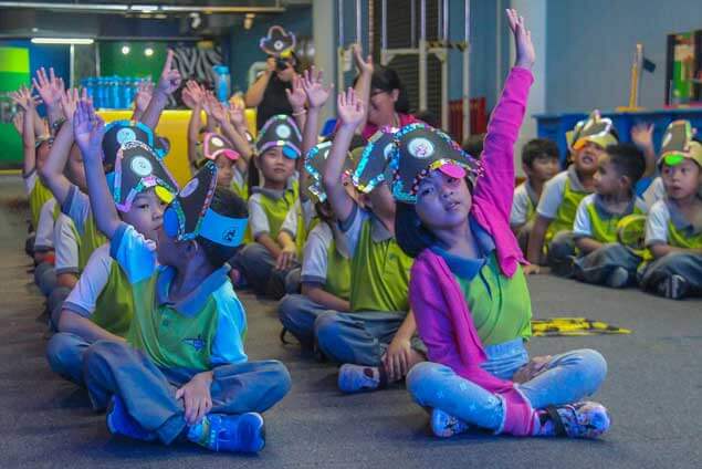 A group of children in matching outfits with decorated hats raise their hands in a classroom setting.
