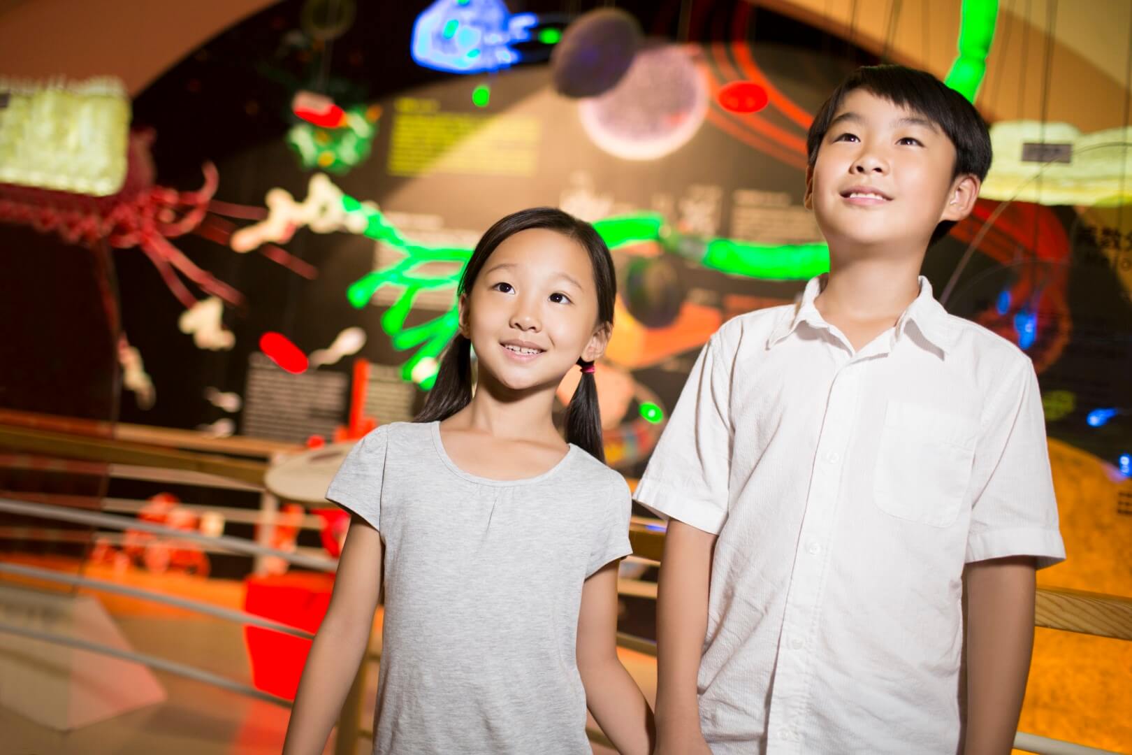 Two smiling children in a science and technology museum looking up at an exhibit.