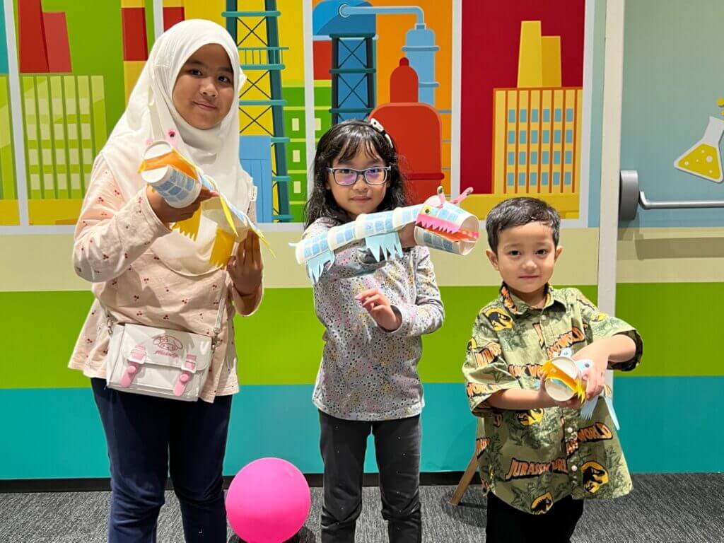 Three smiling children proudly display their handmade paper roll animal crafts against a colorful, cartoon-style cityscape backdrop.