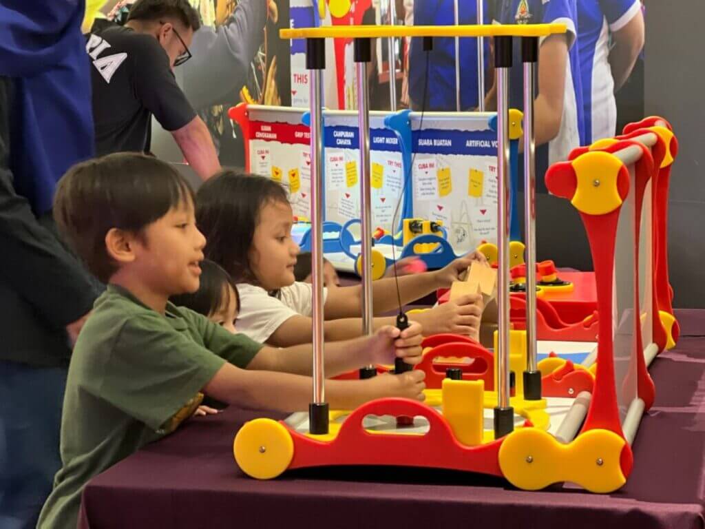 Children interact with a colorful educational exhibit labeled "Grip Test," "Light Mixer," and "Artificial Voice."