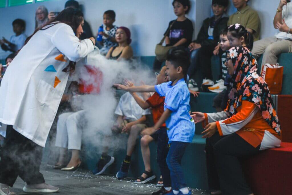 A woman in a lab coat conducts a science experiment with fog in front of a seated crowd of children and adults.