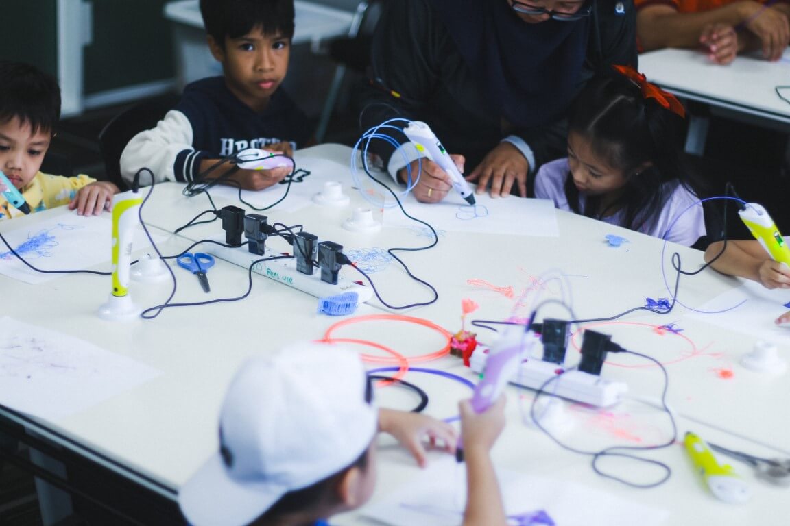Children using 3D printing pens on a white table with an instructor.