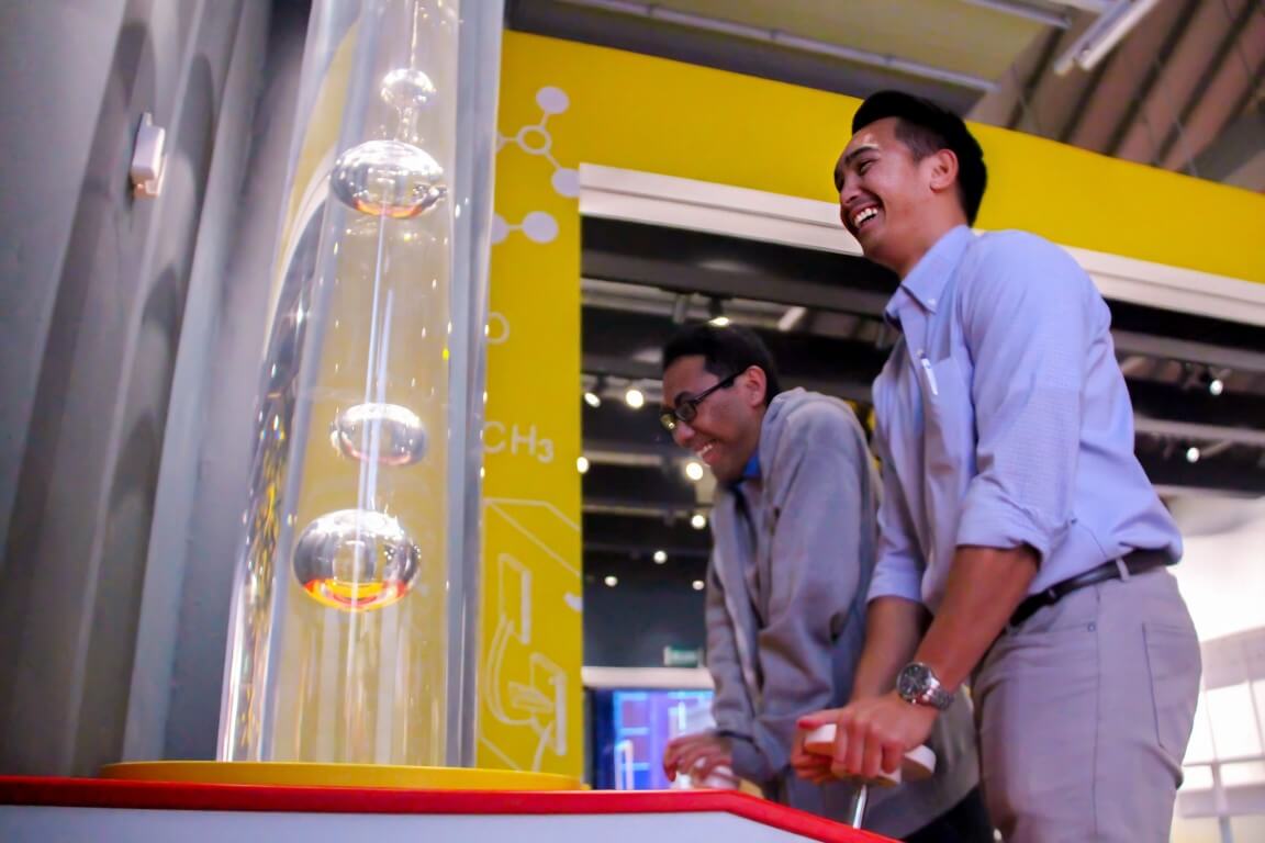 Two men smiling while interacting with a science exhibit featuring rising bubbles in a clear tube.