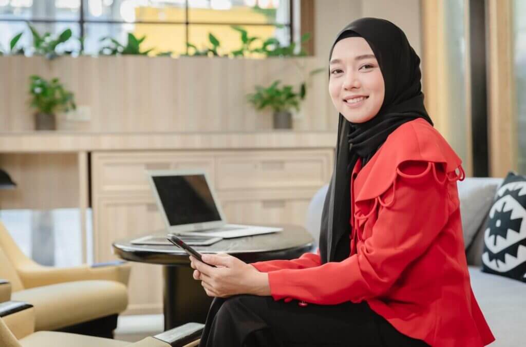 Smiling Muslim businesswoman in hijab with phone, laptop in background.