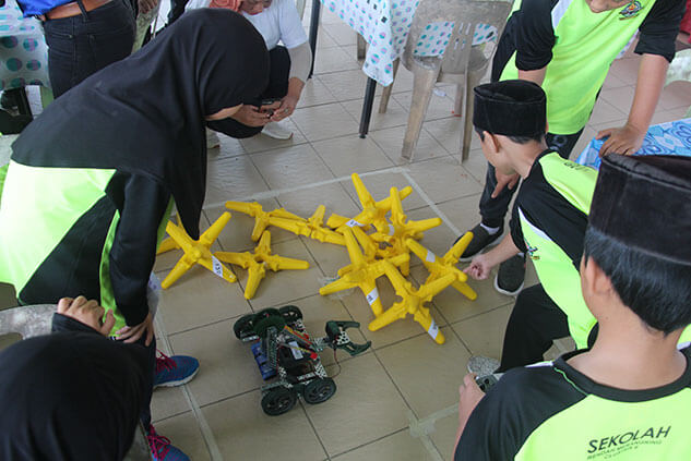 Students gather around a robot and yellow obstacles on a tiled floor during a science activity.