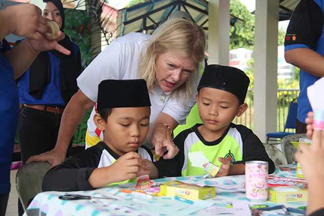 Two young boys in peci hats learn from a woman volunteer during a science or craft activity.