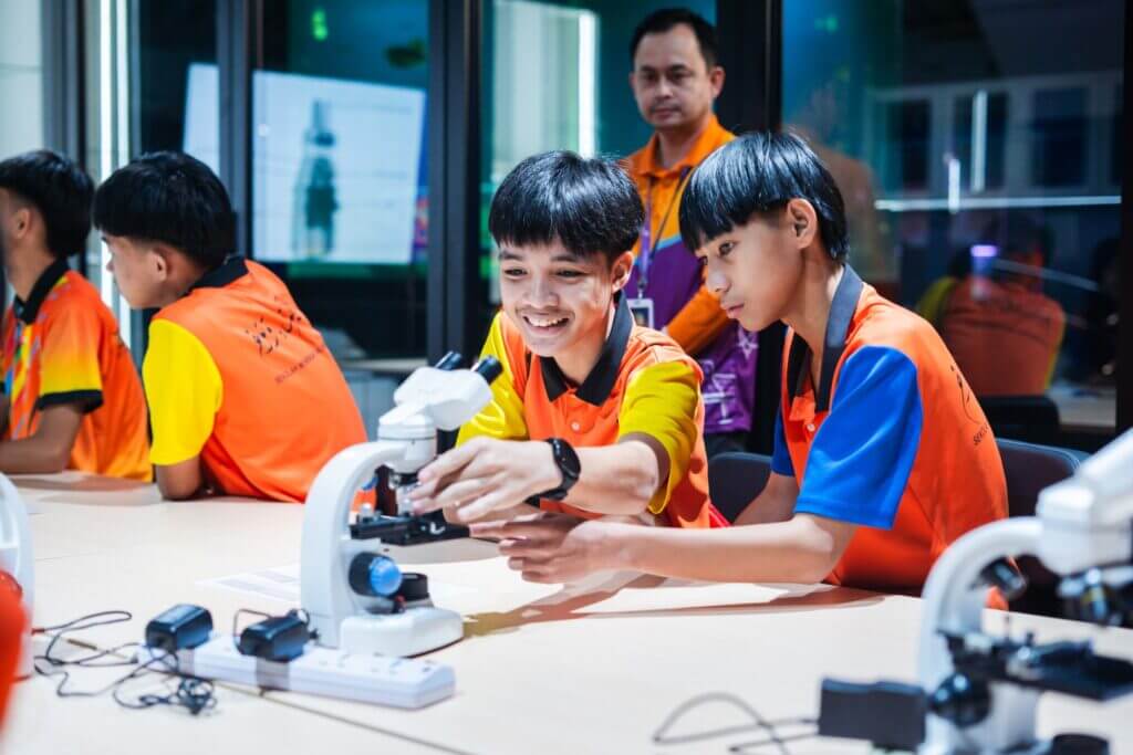 Students in bright shirts examine specimens with a microscope in a science lab.