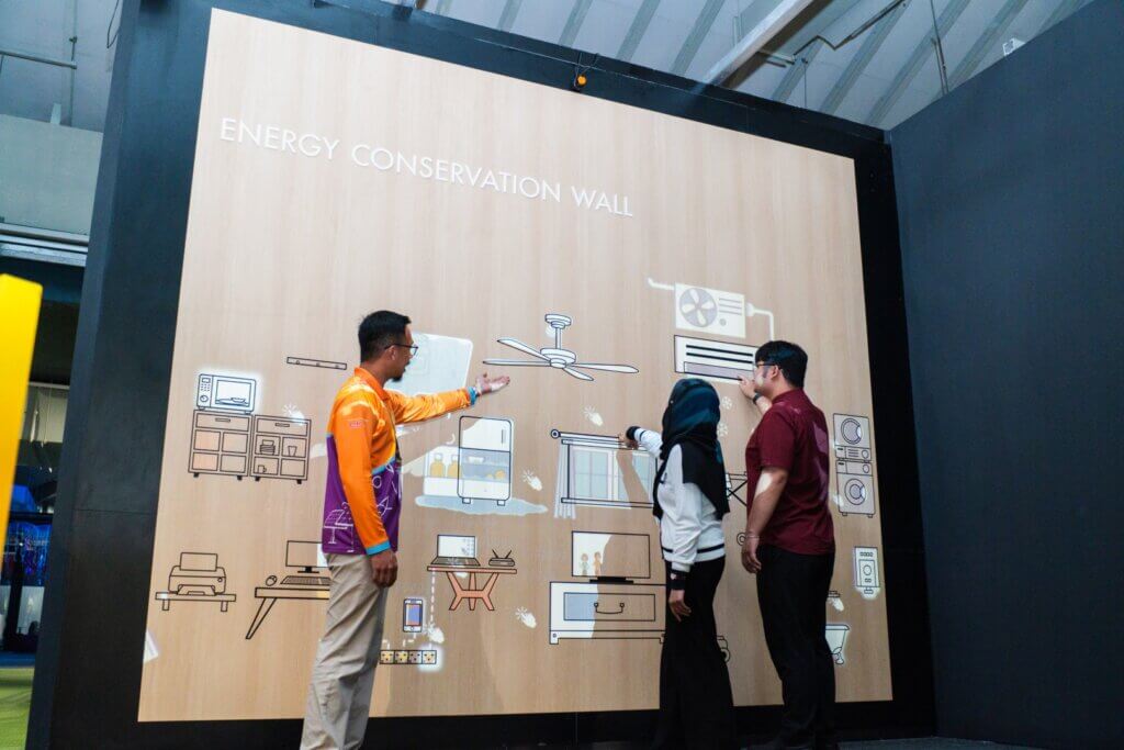 People looking at a wall display titled "Energy Conservation Wall" with illustrations of household appliances.