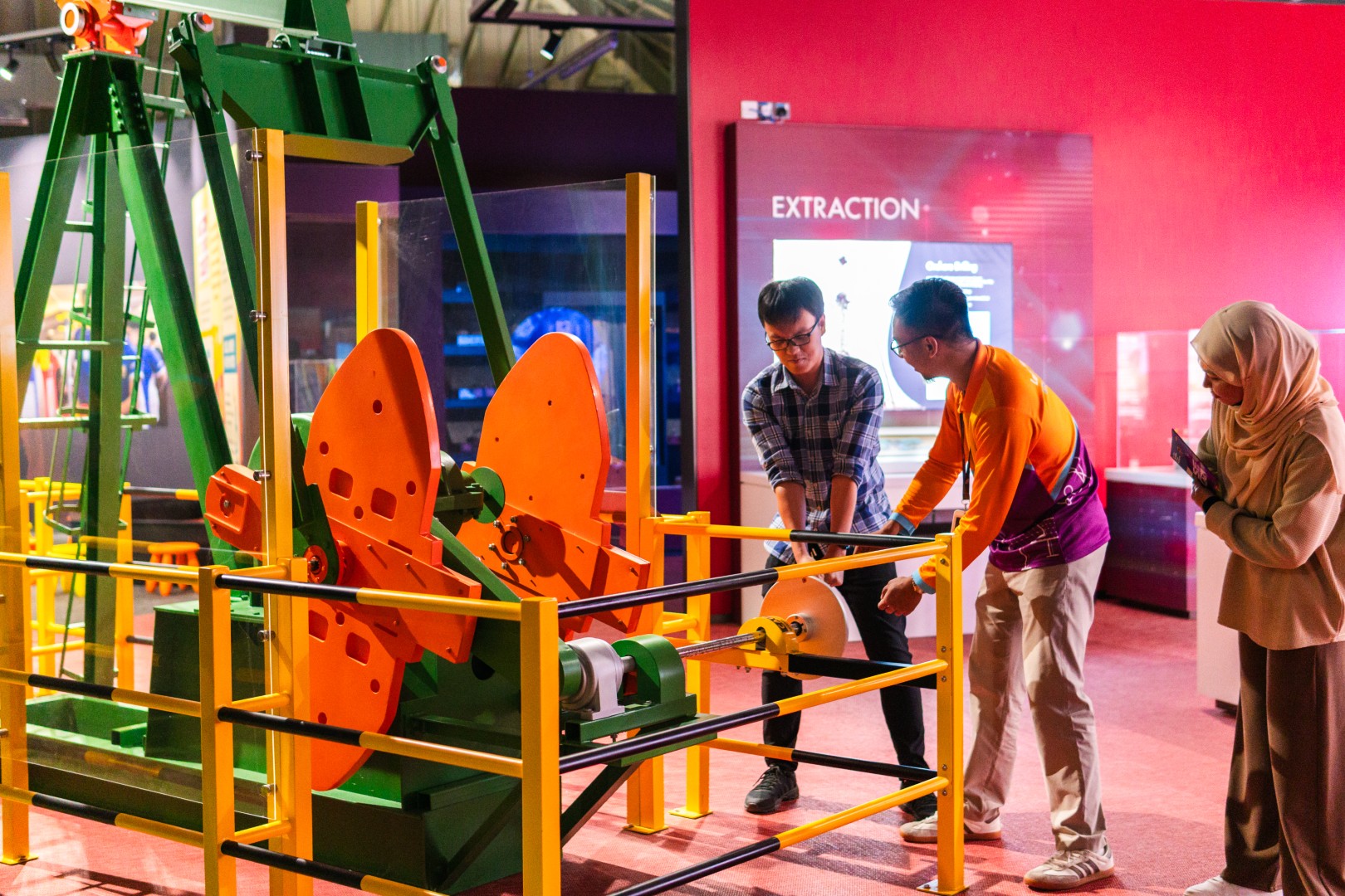 At an exhibit in the "Oil and Gas Zone 8", two people operate a model pump jack while another person looks on.