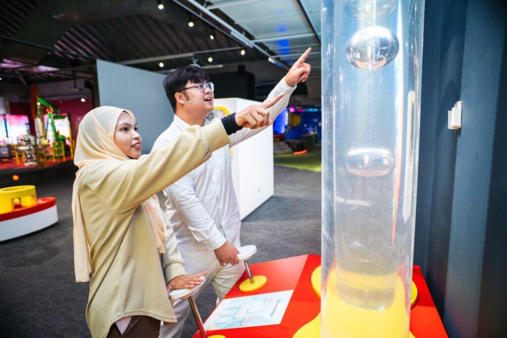 Two people pointing at slow-moving bubbles in a tall tube exhibit.